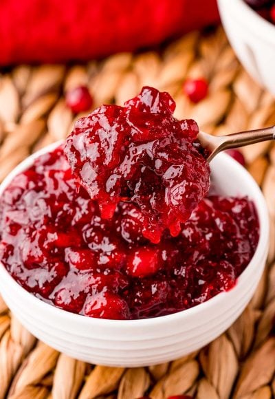 A spoon scooping cranberry sauce out of a white bowl on a wicker placemat.