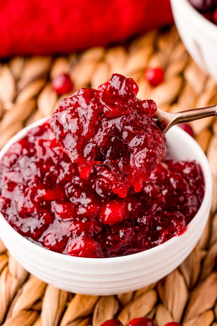 A spoon scooping cranberry sauce out of a white bowl on a wicker placemat.