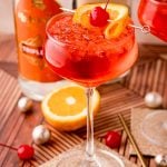 Close up photo of a dirty shirley drink in a cocktail glass on a gold coaster with maraschino cherries, oranges and decorations scattered around.