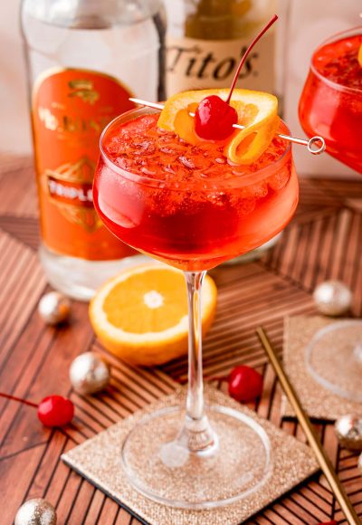 Close up photo of a dirty shirley drink in a cocktail glass on a gold coaster with maraschino cherries, oranges and decorations scattered around.