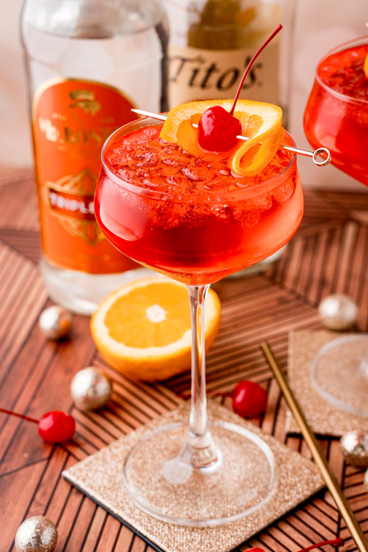 Close up photo of a dirty shirley drink in a cocktail glass on a gold coaster with maraschino cherries, oranges and decorations scattered around.