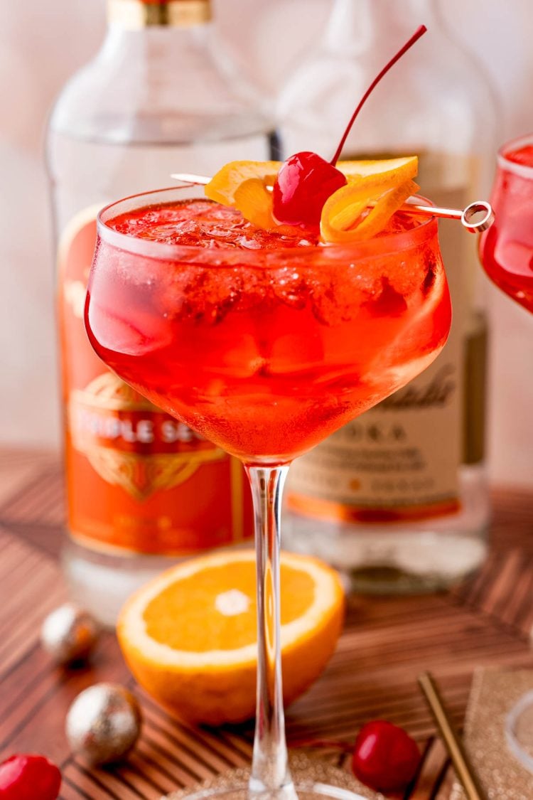 Close up photo of a dirty shirley drink in a cocktail glass on a gold coaster with maraschino cherries, oranges and decorations scattered around.