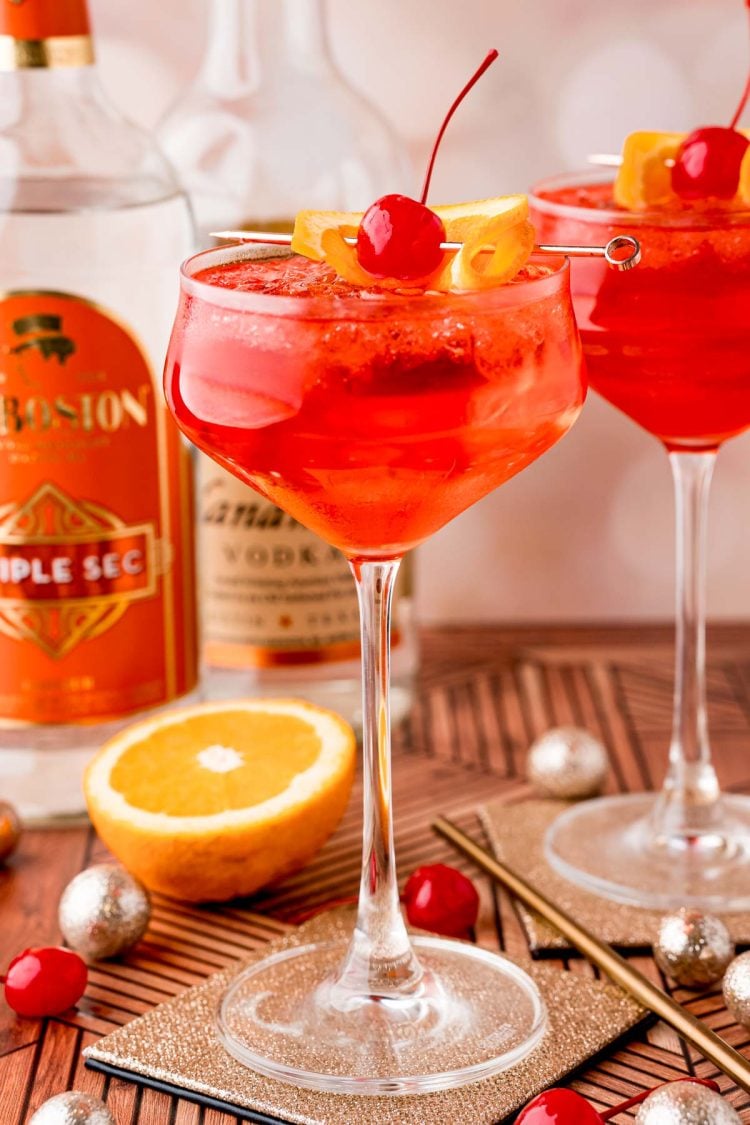 Close up photo of a dirty shirley drink in a cocktail glass on a gold coaster with maraschino cherries, oranges and decorations scattered around.