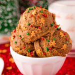 Close up photo of a white bowl filled with edible gingerbread cookies dough on a red sparkly placemat with a glass of milk in the background.