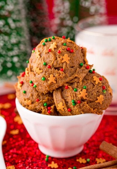 Close up photo of a white bowl filled with edible gingerbread cookies dough on a red sparkly placemat with a glass of milk in the background.