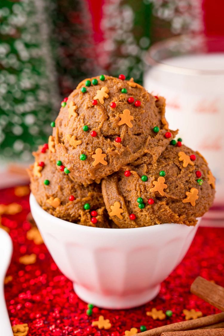 Close up photo of a white bowl filled with edible gingerbread cookies dough on a red sparkly placemat with a glass of milk in the background.