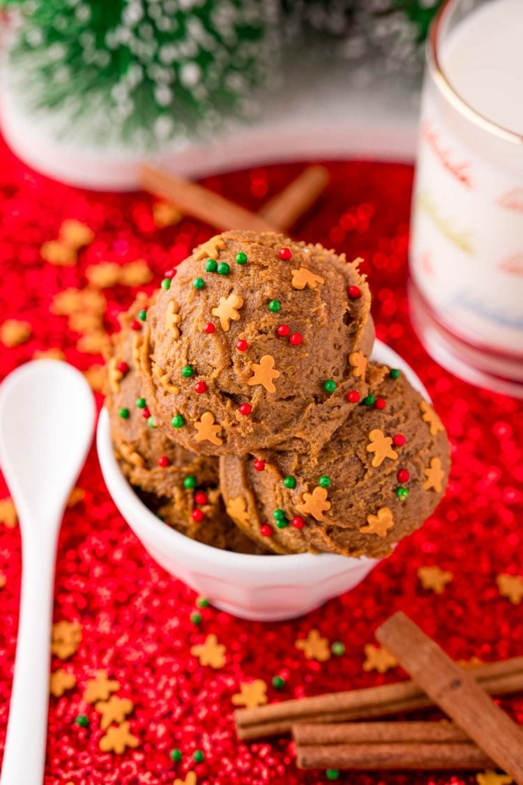A bowl of edible gingerbread cookie dough on a red surface with cinnamon sticks and a glass of milk next to it.
