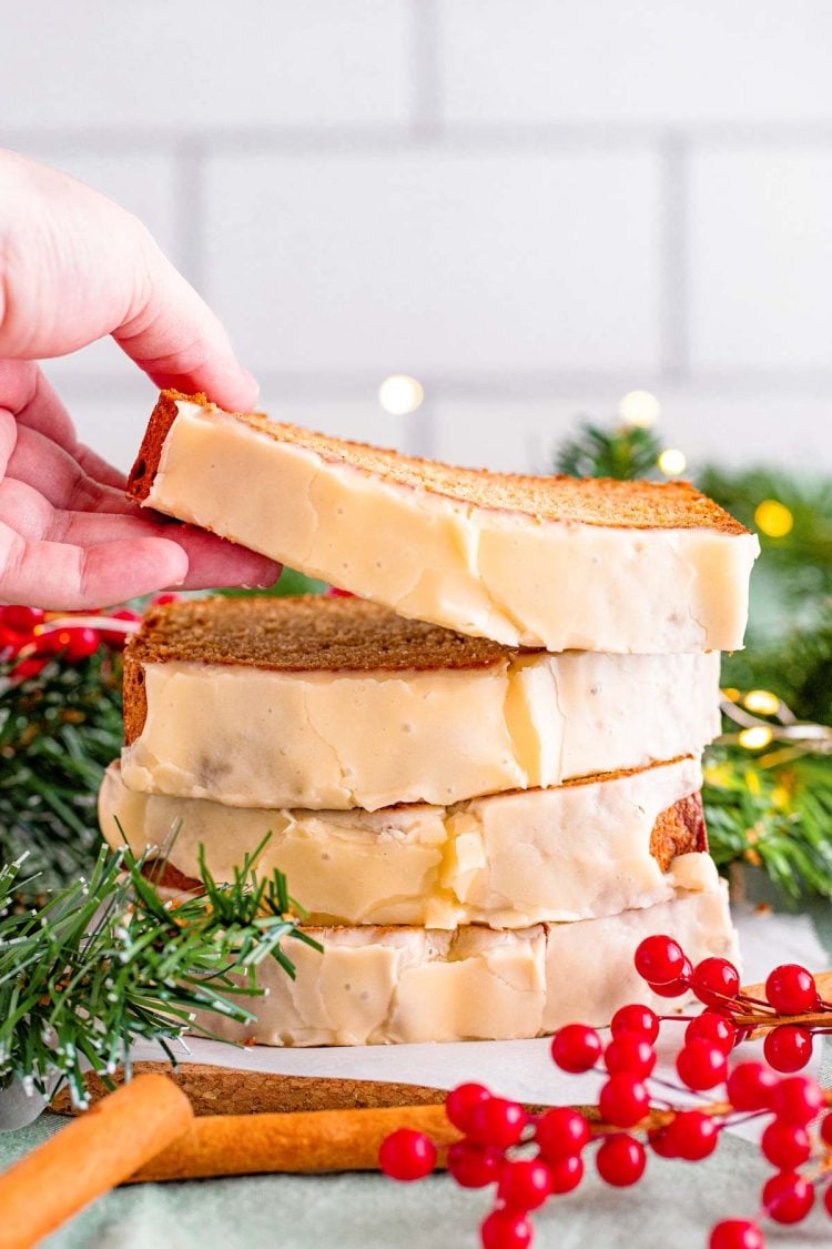 Close up photo of a stack of eggnog bread slices, a woman's hand is taking the top slice off. Holiday decor surrounds the bread.