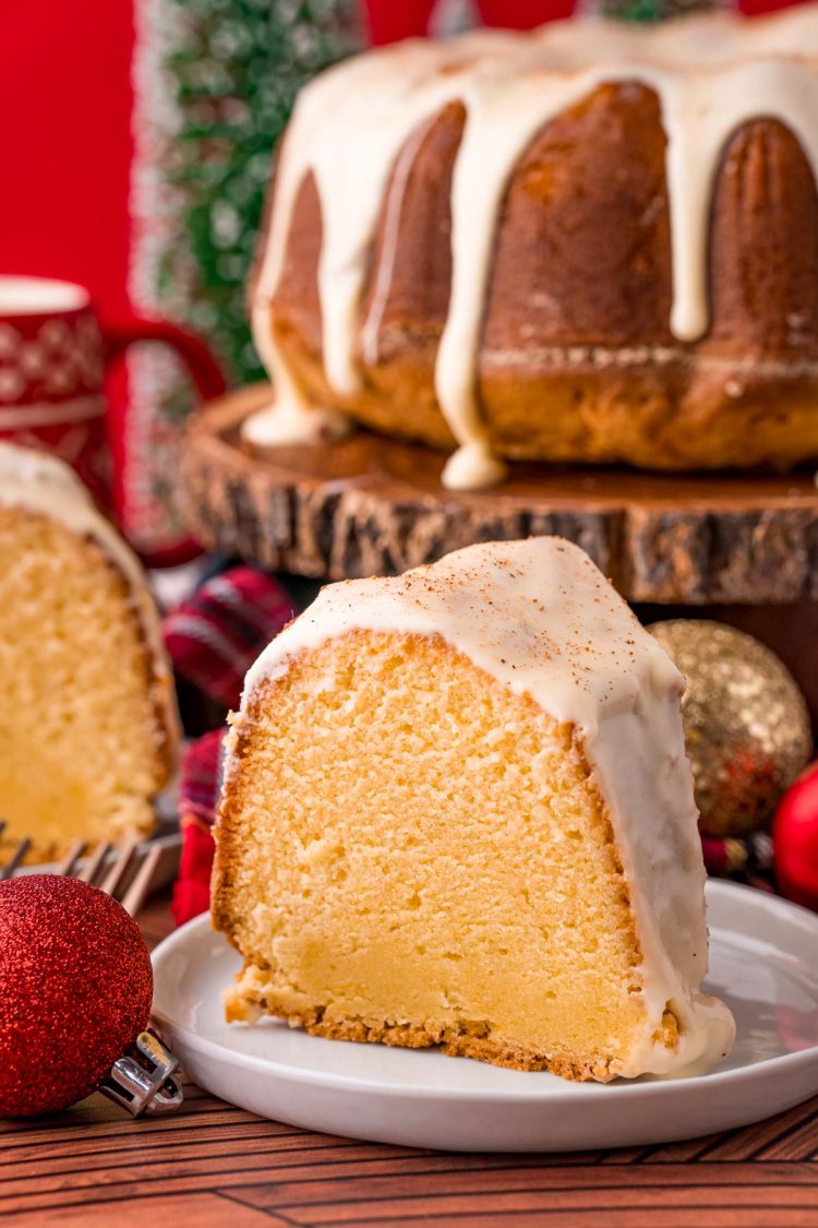 Close up photo of a slice of eggnog pound cake on a white plate with the rest of the cake in the background decorated for the holidays.