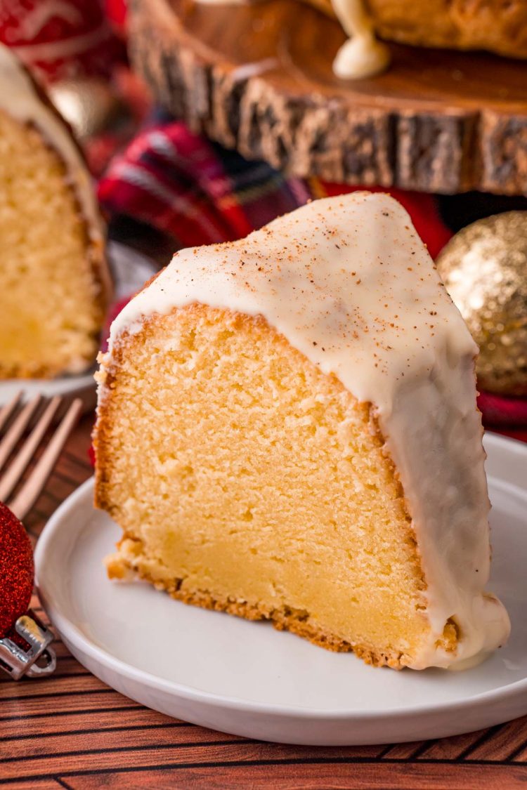 Close up photo of a slice of eggnog pound cake on a white plate with the rest of the cake in the background decorated for the holidays.