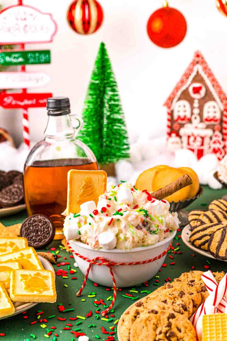 Close up photo of a white bowl filled with christmas dessert dip with cookies around it on a green surface.