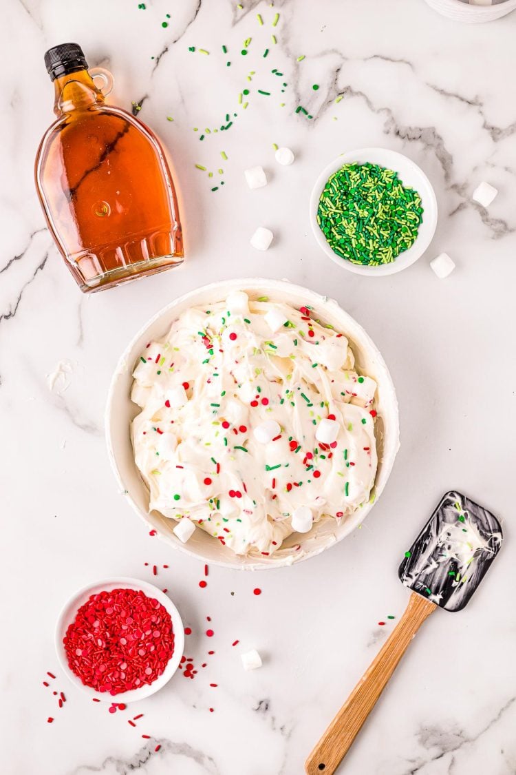 Overhead photo of christmas dessert dip in a white bowl on a marble surface with a rubber spatula, maple syrup, and bowls of sprinkles around it.