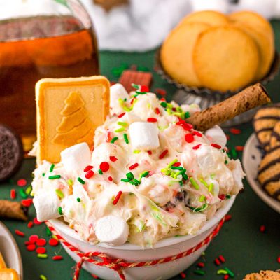 Close up photo of a white bowl filled with christmas dessert dip with cookies around it on a green surface.