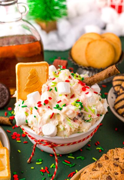 Close up photo of a white bowl filled with christmas dessert dip with cookies around it on a green surface.