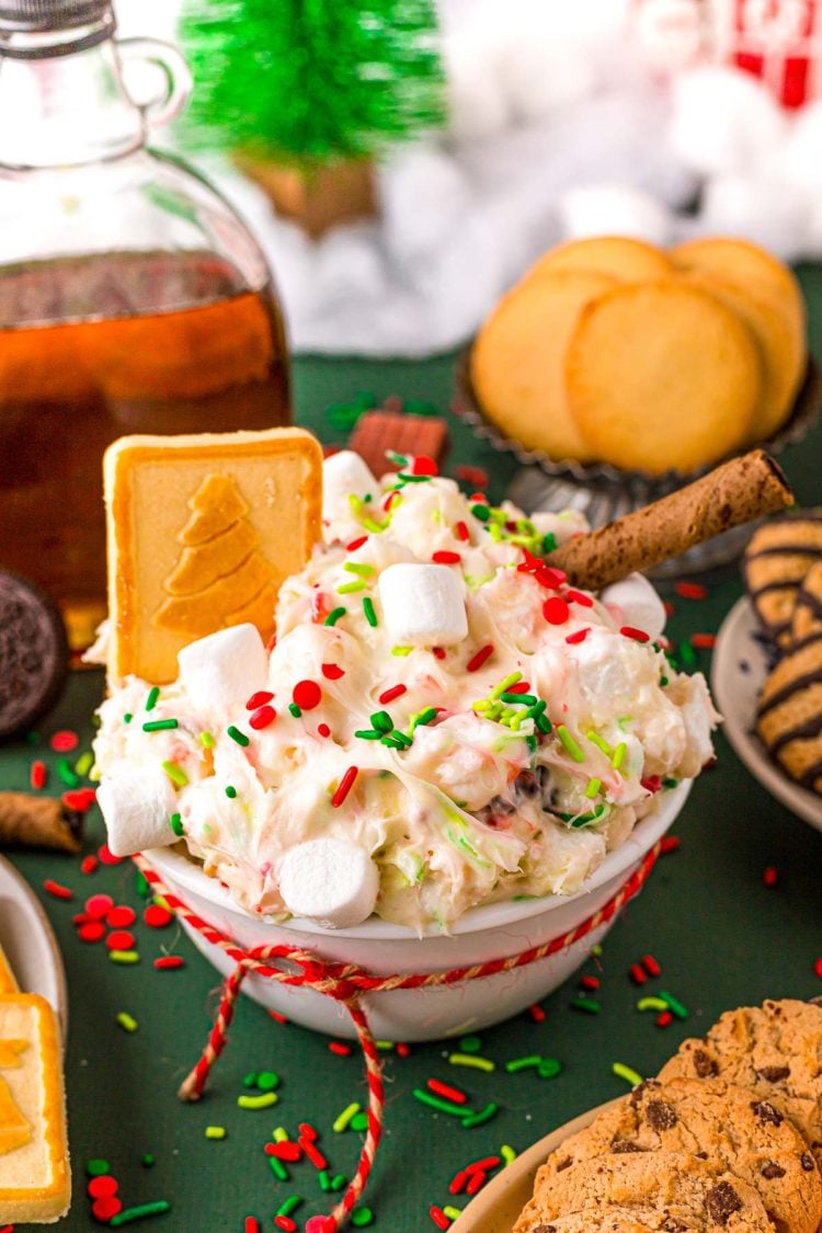 Close up photo of a white bowl filled with christmas dessert dip with cookies around it on a green surface.