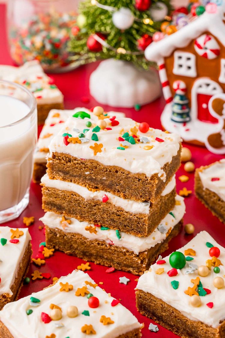 Angled photo of a stack of three gingerbread cookies bars on a red table with more bars and holiday decorations scattered around.