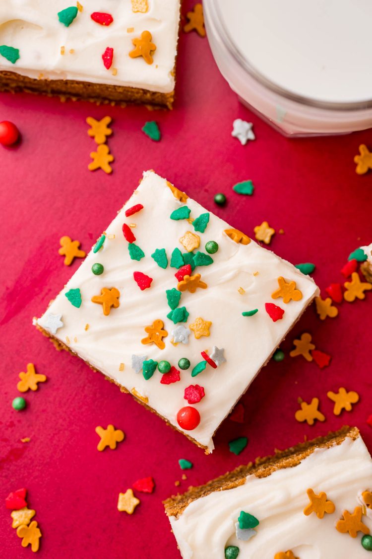 A gingerbread cookie bar with cream cheese frosting on a red table with other bars, sprinkles, and a glass of milk around it.
