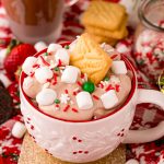Close up photo of a white holiday mug filled with hot chocolate dip with marshmallows and sprinkles on top. A shortbread cookie is stuck in the top of the dip.