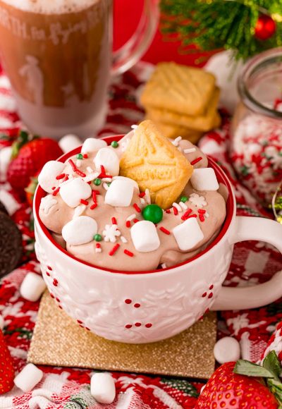 Close up photo of a white holiday mug filled with hot chocolate dip with marshmallows and sprinkles on top. A shortbread cookie is stuck in the top of the dip.