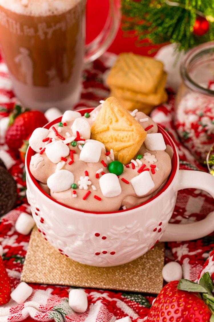 Close up photo of a white holiday mug filled with hot chocolate dip with marshmallows and sprinkles on top. A shortbread cookie is stuck in the top of the dip.