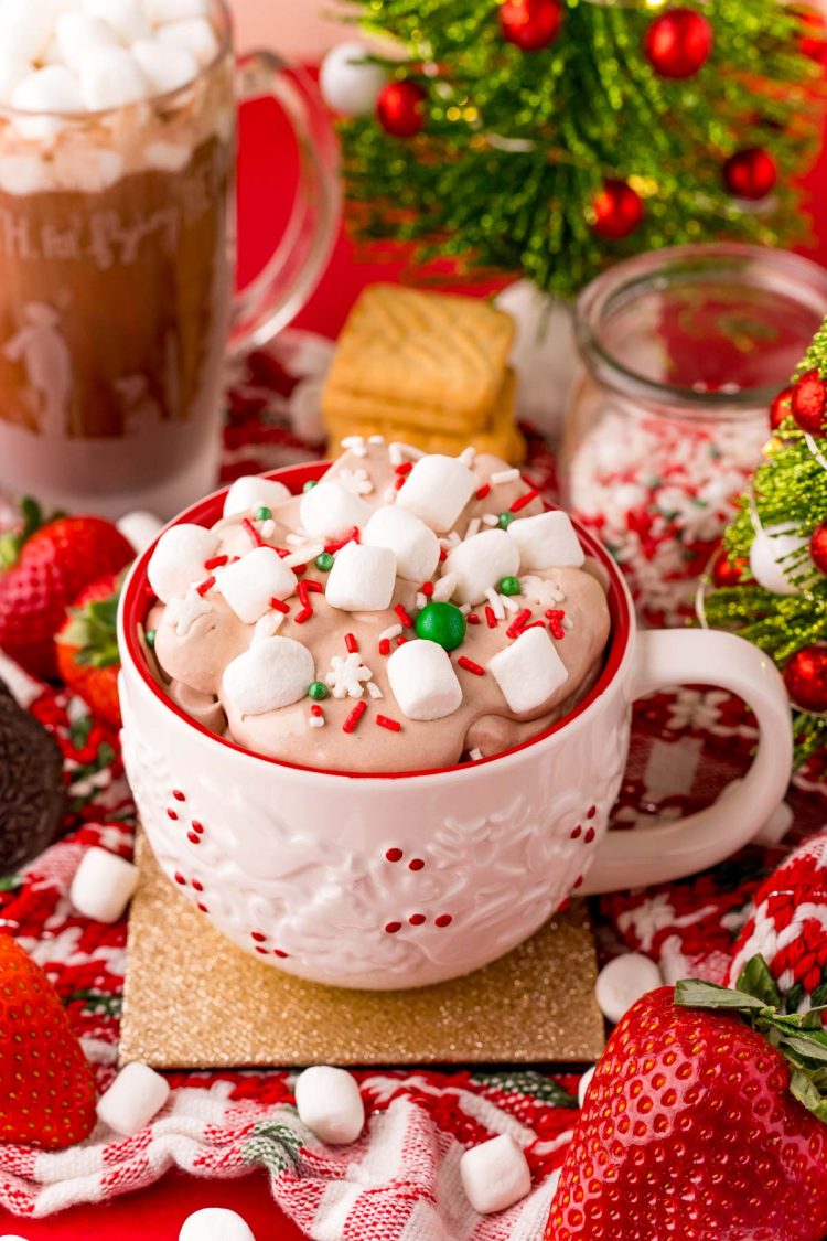 Close up photo of a white holiday mug filled with hot chocolate dip with marshmallows and sprinkles on top. 