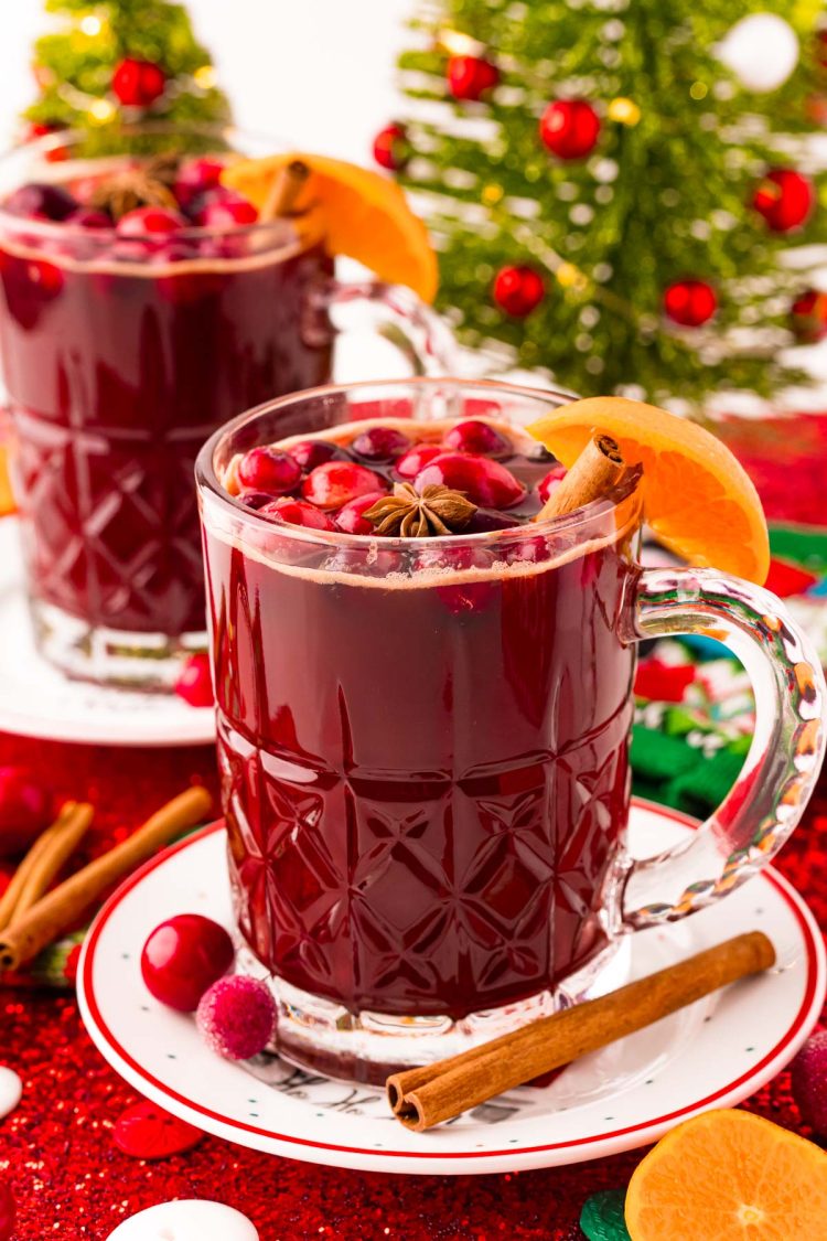 Two mugs of Kinderpunsch on saucers with cranberries, oranges, and cinnamon sticks scattered around. Holiday decor in the background.