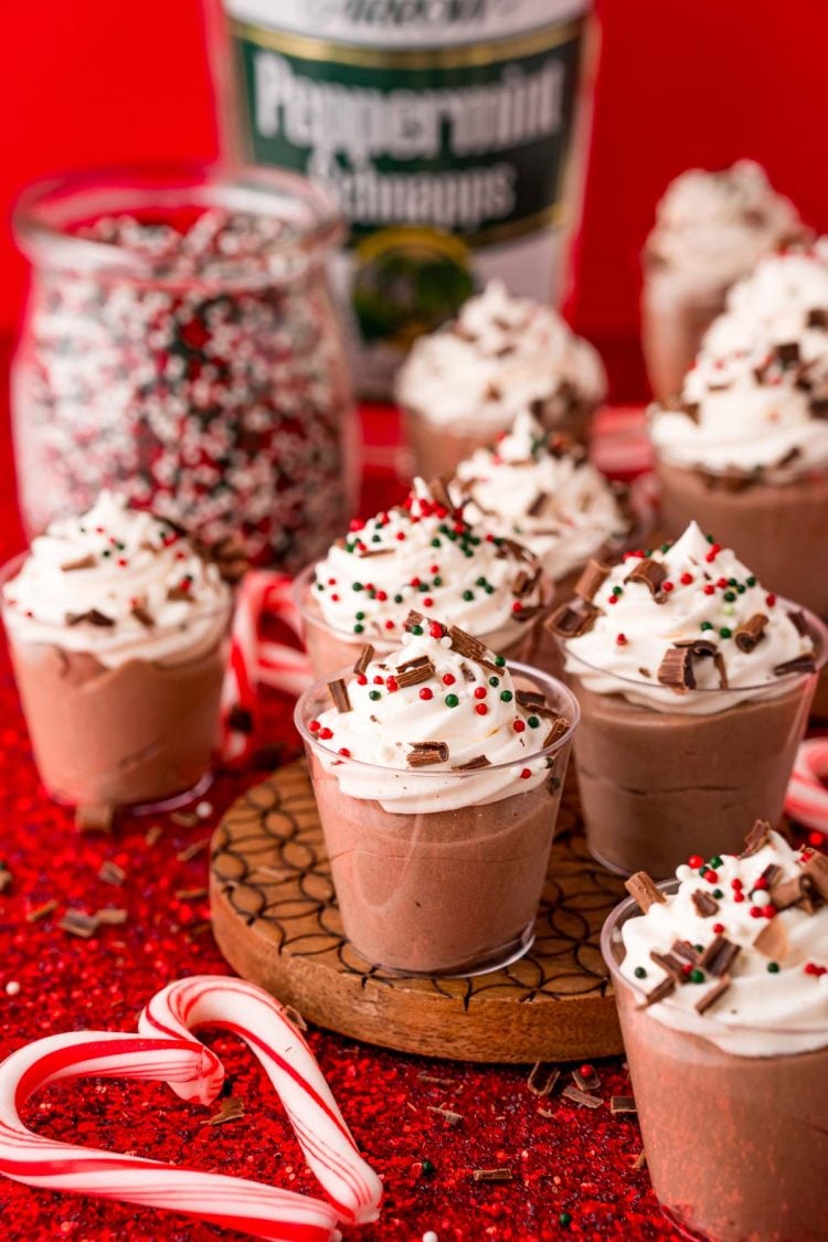 Peppermint chocolate pudding shots on a red table with mini candy canes and a bottle of sprinkles and peppermint schnapps in the background.