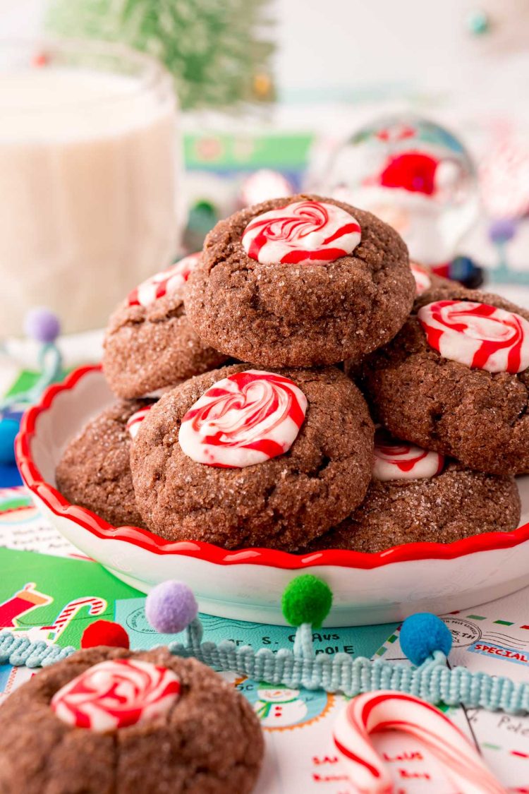 Chocolate peppermint thumbprint cookies on a white plate with a red rim on christmas wrapping paper with a glass of milk in the background.