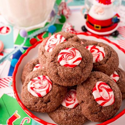 Close up photo of a small plate filled with chocolate peppermint thumbprint cookies. Holiday decor and a glass of milk are around the plate.