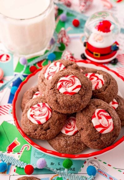 Close up photo of a small plate filled with chocolate peppermint thumbprint cookies. Holiday decor and a glass of milk are around the plate.