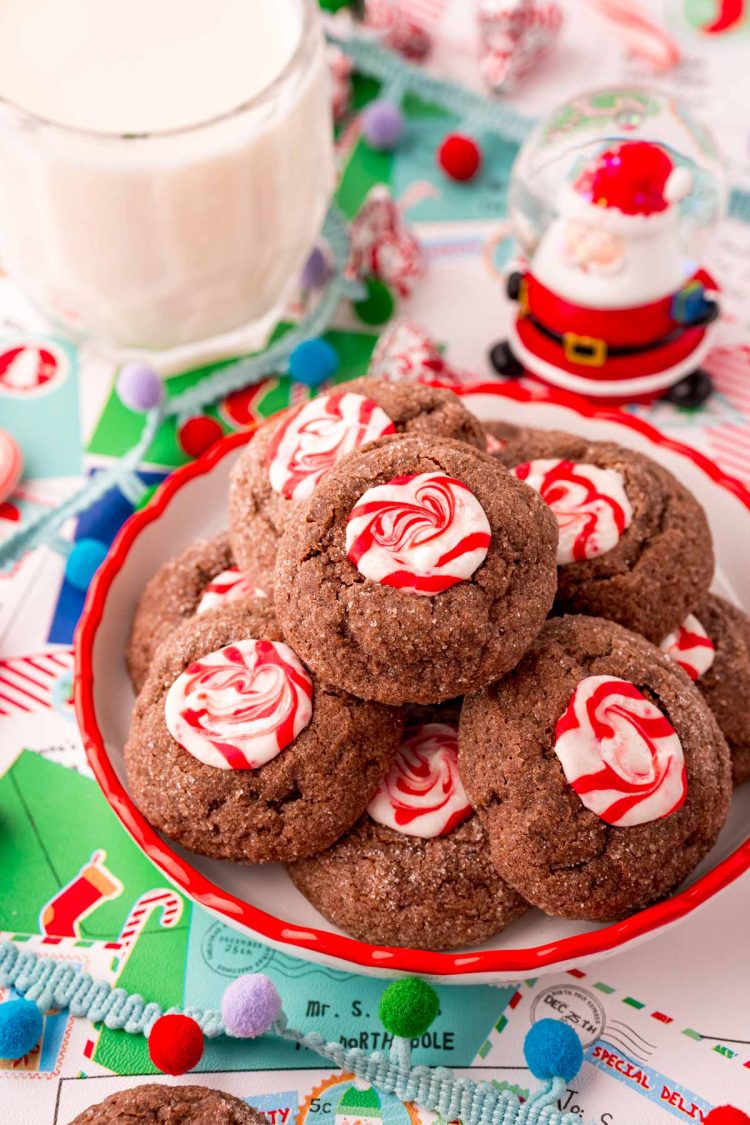 Close up photo of a small plate filled with chocolate peppermint thumbprint cookies. Holiday decor and a glass of milk are around the plate.