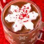 Close up photo of a mug of peppermint hot chocolate on a red surface with candy canes and chocolate and red ribbon around it.