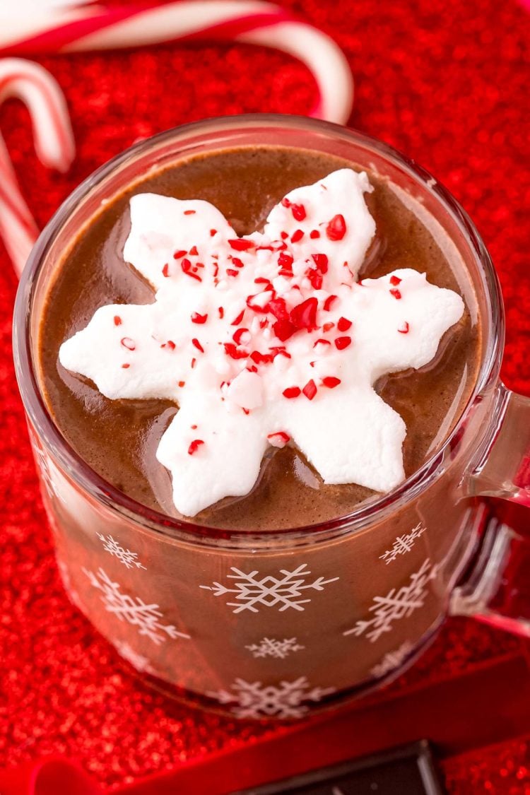 Close up photo of a mug of peppermint hot chocolate on a red surface with candy canes and chocolate and red ribbon around it.