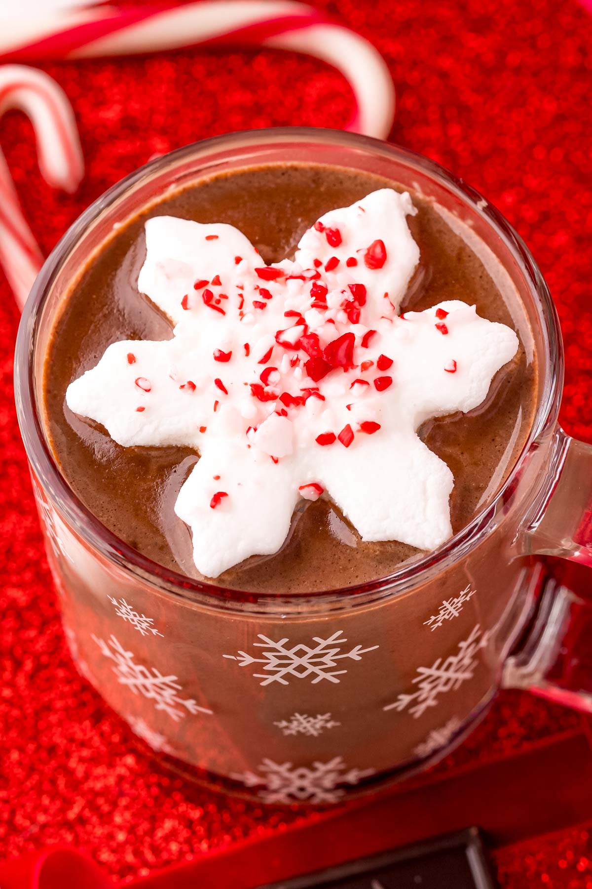 Close up photo of a mug of peppermint hot chocolate on a red surface with candy canes and chocolate and red ribbon around it.