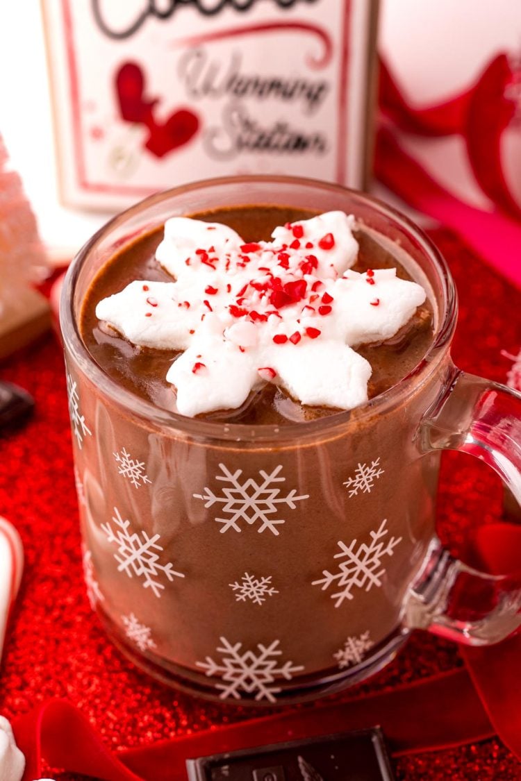 Close up photo of a mug of peppermint hot chocolate on a red surface with candy canes and chocolate and red ribbon around it.