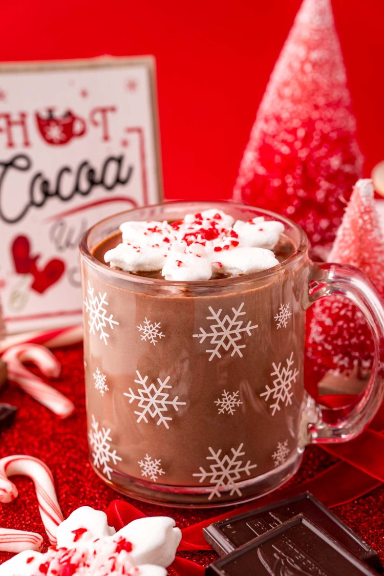 Close up photo of a glass mug with white snowflakes on it filled with peppermint hot chocolate and topped with a snowflake shaped marshmallow.