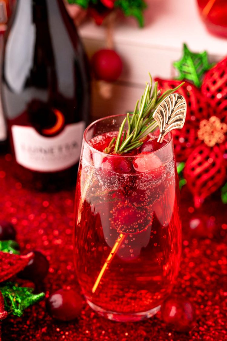 Close up photo of a stemless champagne glass with a poinsettia cocktail (cranberry mimosa) in it with a bottle of prosecco and holiday decor in the background.