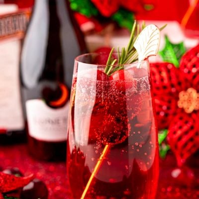 Close up photo of a stemless champagne glass with a poinsettia cocktail (cranberry mimosa) in it with a bottle of prosecco and holiday decor in the background.