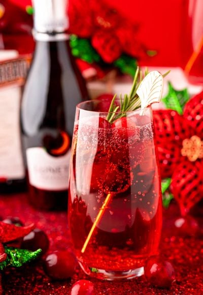Close up photo of a stemless champagne glass with a poinsettia cocktail (cranberry mimosa) in it with a bottle of prosecco and holiday decor in the background.
