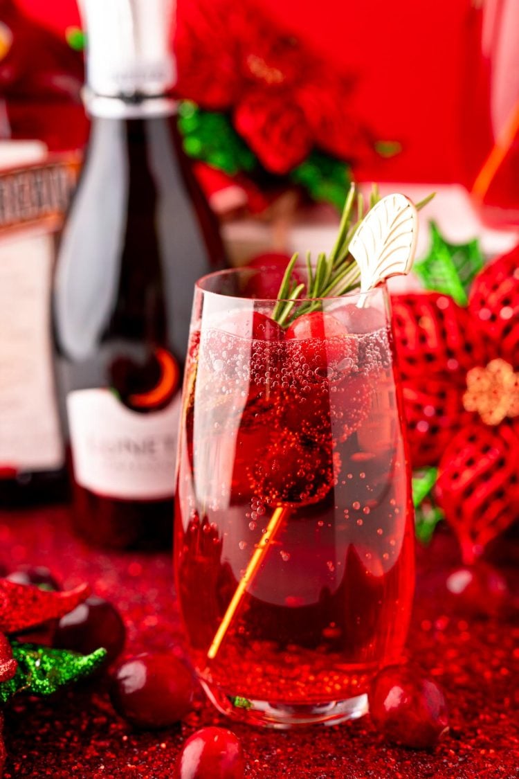 Close up photo of a stemless champagne glass with a poinsettia cocktail (cranberry mimosa) in it with a bottle of prosecco and holiday decor in the background.