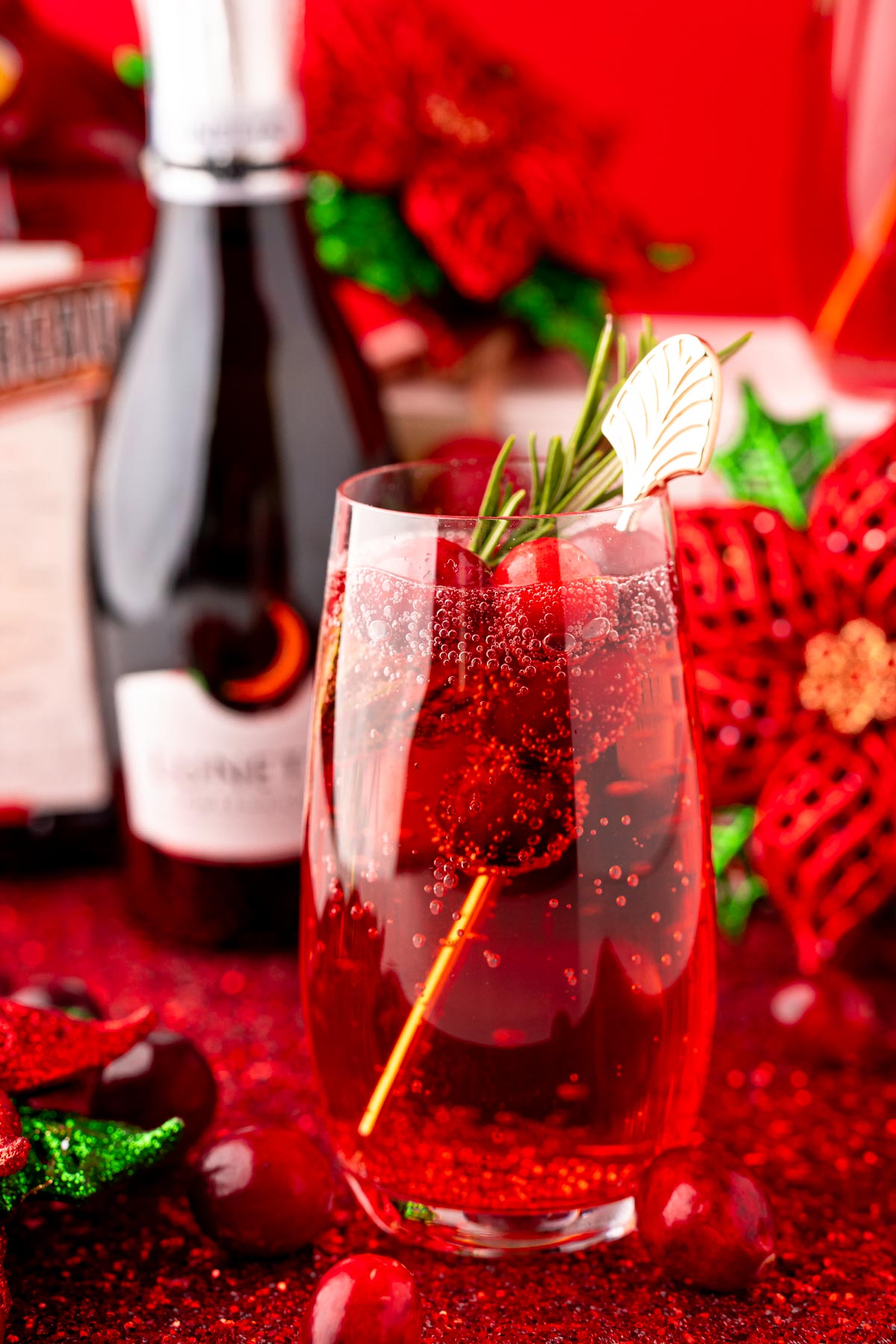 Close up photo of a stemless champagne glass with a poinsettia cocktail (cranberry mimosa) in it with a bottle of prosecco and holiday decor in the background.