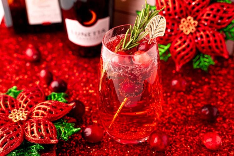 Close up photo of a stemless champagne glass with a poinsettia cocktail (cranberry mimosa) in it with a bottle of prosecco and holiday decor in the background.