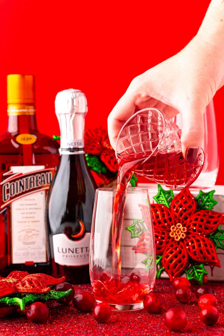 Cranberry juice being poured into a stemless champagne flute from a shot glass.