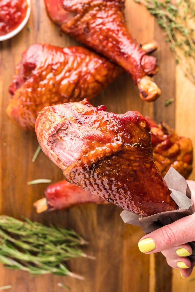 Overhead photo of a woman's hand holding a turkey leg with a bite missing from it over a cutting board with more turkey legs on it.