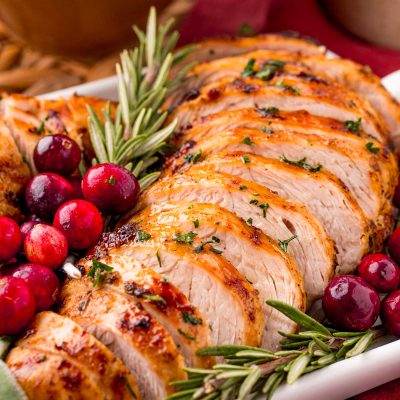 Close up photo of turkey tenderloin on a serving tray with herbs and cranberries on a red napkin with a bowl of mashed potatoes in the background.