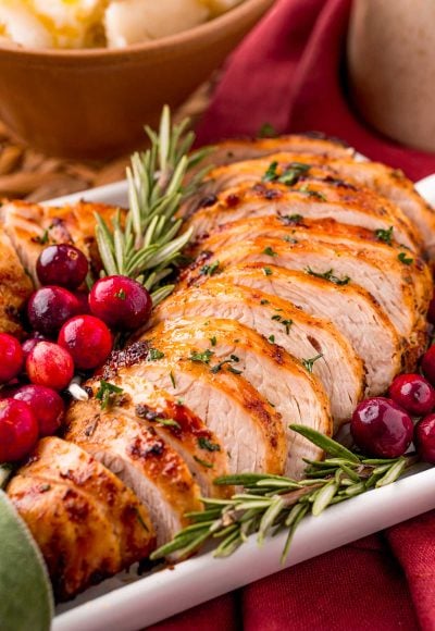 Close up photo of turkey tenderloin on a serving tray with herbs and cranberries on a red napkin with a bowl of mashed potatoes in the background.