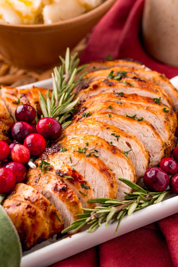 Close up photo of turkey tenderloin on a serving tray with herbs and cranberries on a red napkin with a bowl of mashed potatoes in the background.