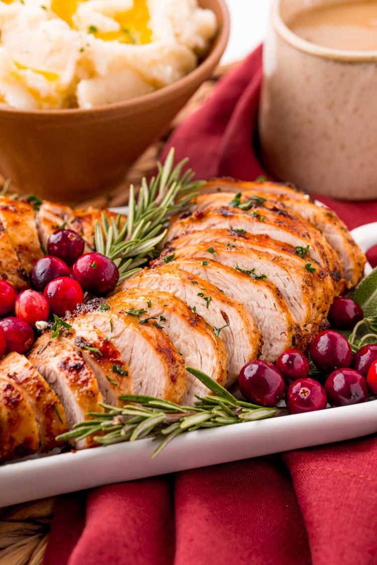 Close up photo of turkey tenderloin on a serving tray with herbs and cranberries on a red napkin with a bowl of mashed potatoes in the background.