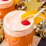 A close up photo of party punch garnished with a raspberry and orange slice on a gold coaster with party decorations in the background.