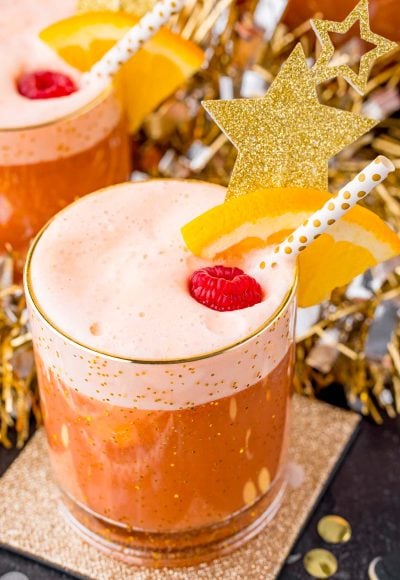 A close up photo of party punch garnished with a raspberry and orange slice on a gold coaster with party decorations in the background.